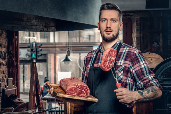 The chef, in the kitchen, holds a dry-aged ribeye steak with a fork in one hand, and a wooden cutting board with the aging steak on it in the other.