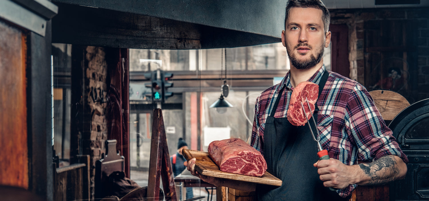 In the kitchen, a chef is poised over a wooden cutting board, a perfectly marbled dry-aged ribeye steak resting upon it. His left hand steadies the meat with a fork, as he prepares to transform the raw cut into a culinary masterpiece.